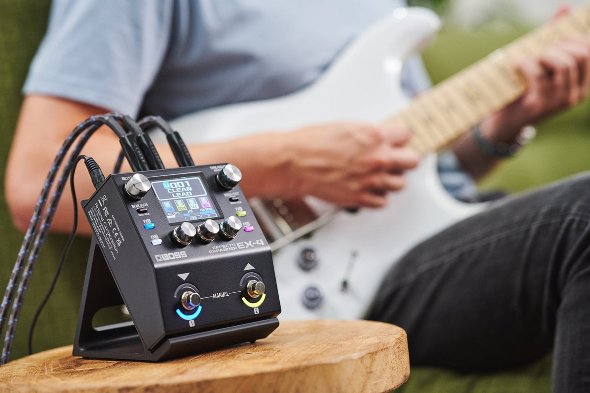 The BOSS EX-4 Effects Expander on a wooden stool beside a guitarist playing a white electric guitar