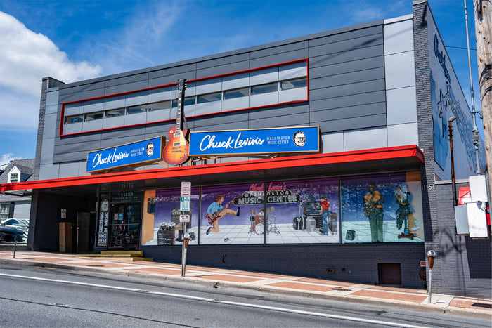 A street-facing view of a modern gray building with large Chuck Lewis's signage, a red awning, and a guitar decoration outside. The storefront windows show colorful winter sports murals, and the scene is set against a blue sky with white clouds.